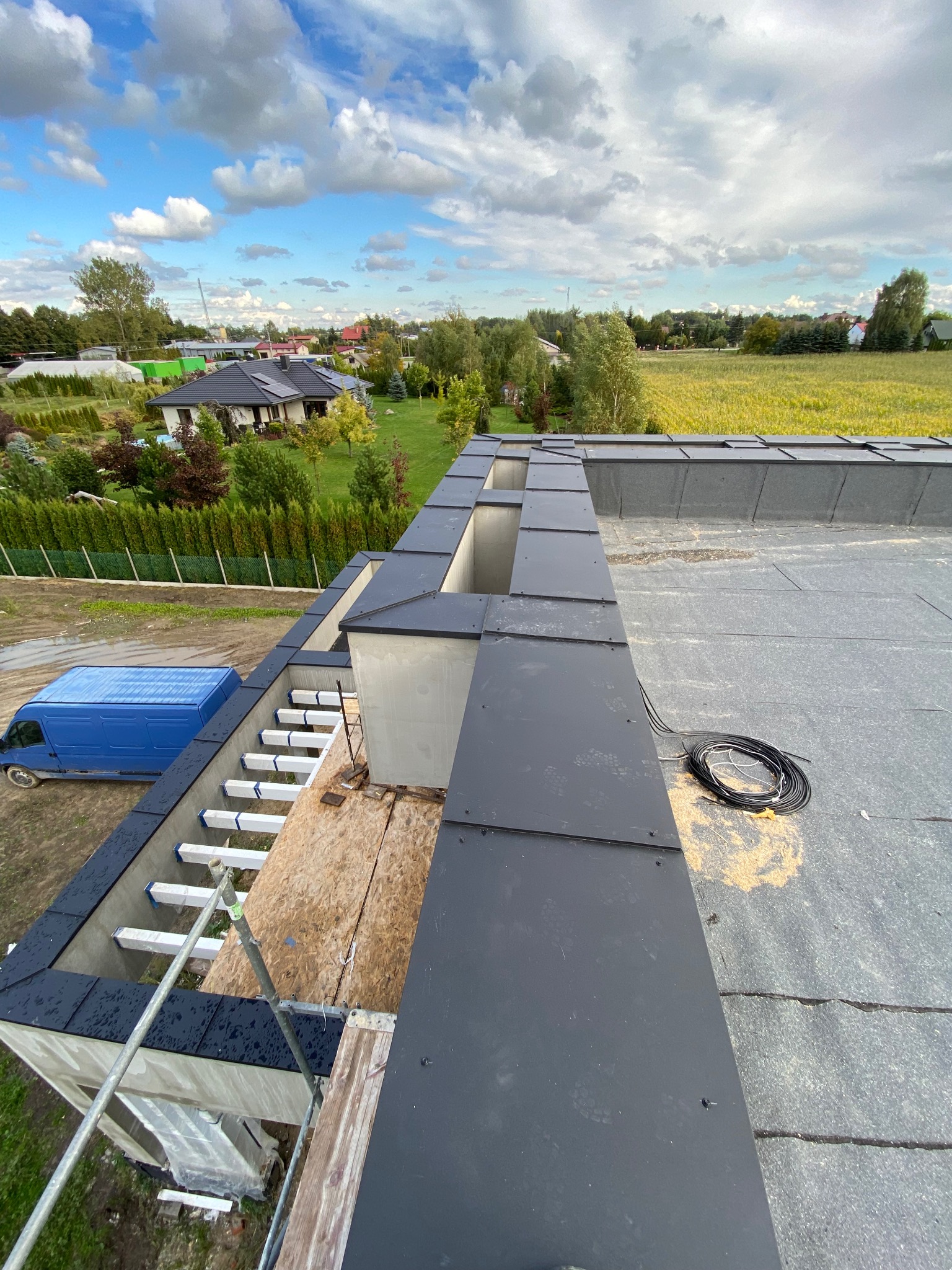 Częściowy widok dachu z blachy na tle błękitnego nieba z chmurami. Widoczny fragment konstrukcji, kabel i niebieski samochód dostawczy. Prace dekarskie w toku.