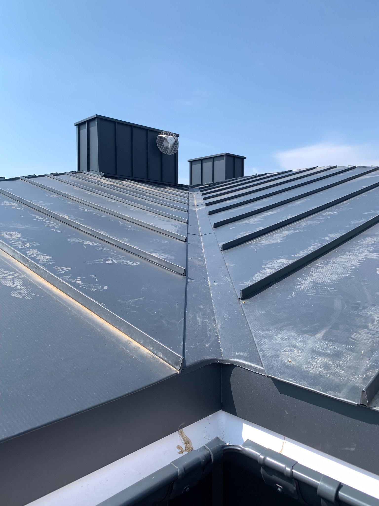 Szary dach z blachy trapezowej z kominami i zabezpieczeniem komina, widok z góry w słoneczny dzień. Widoczne ślady po butach na dachu.