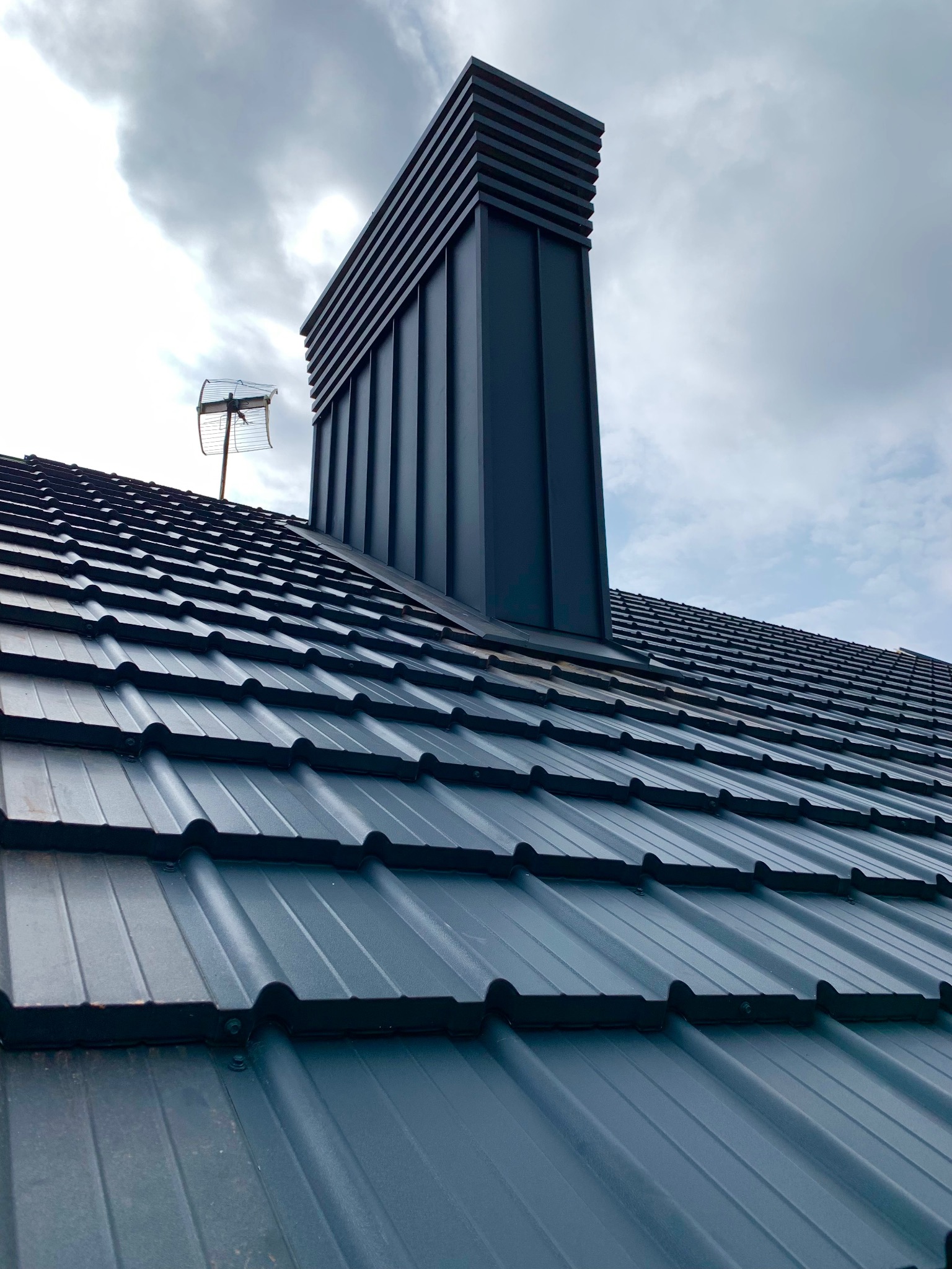 Grafitowy dach z blachy trapezowej z nowoczesnym kominem wentylacyjnym i anteną telewizyjną na tle pochmurnego nieba. Widok z dołu.