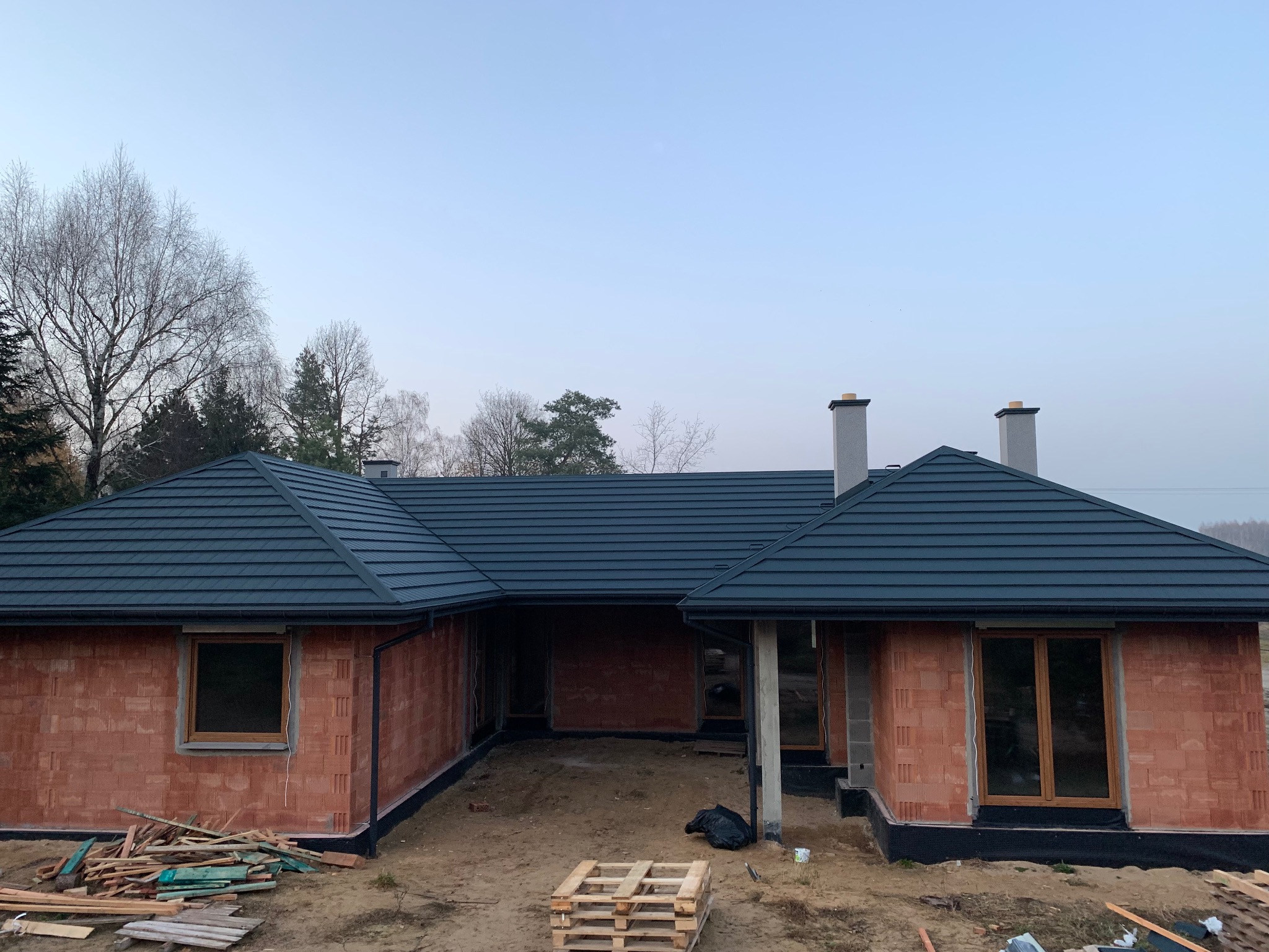 Dom w trakcie budowy z ciemnym dachem z dachówki ceramicznej i kominami. Widoczne ściany z czerwonej cegły i elementy konstrukcyjne.