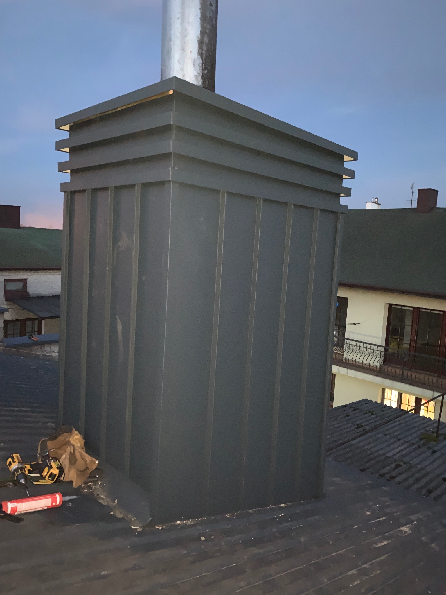 Szary, metalowy komin z narzędziami na dachu z blachy falistej, widziany z dołu pod kątem. W tle budynki mieszkalne i wieczorne niebo.