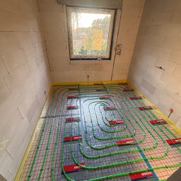 HydroSpec Jacek Wieczorek - Układanie zielonych rur ogrzewania podłogowego na folii aluminiowej w surowym pomieszczeniu z oknem. Widoczne mocowania rur i fragment ściany z betonu komórkowego.