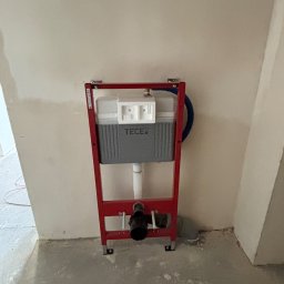 HydroSpec Jacek Wieczorek - Stelaż podtynkowy WC marki TECE z czerwoną ramą, zamontowany na surowej ścianie w trakcie prac wykończeniowych. Widoczne elementy instalacji hydraulicznej.