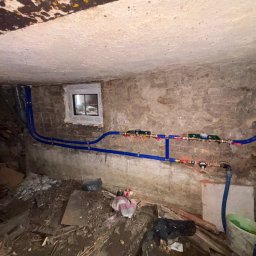 HydroSpec Jacek Wieczorek - Niebieskie rury instalacji wodnej z zaworami zamontowane w surowym, ceglanym pomieszczeniu z małym oknem. Widoczne ślady remontu i gruz na podłodze.