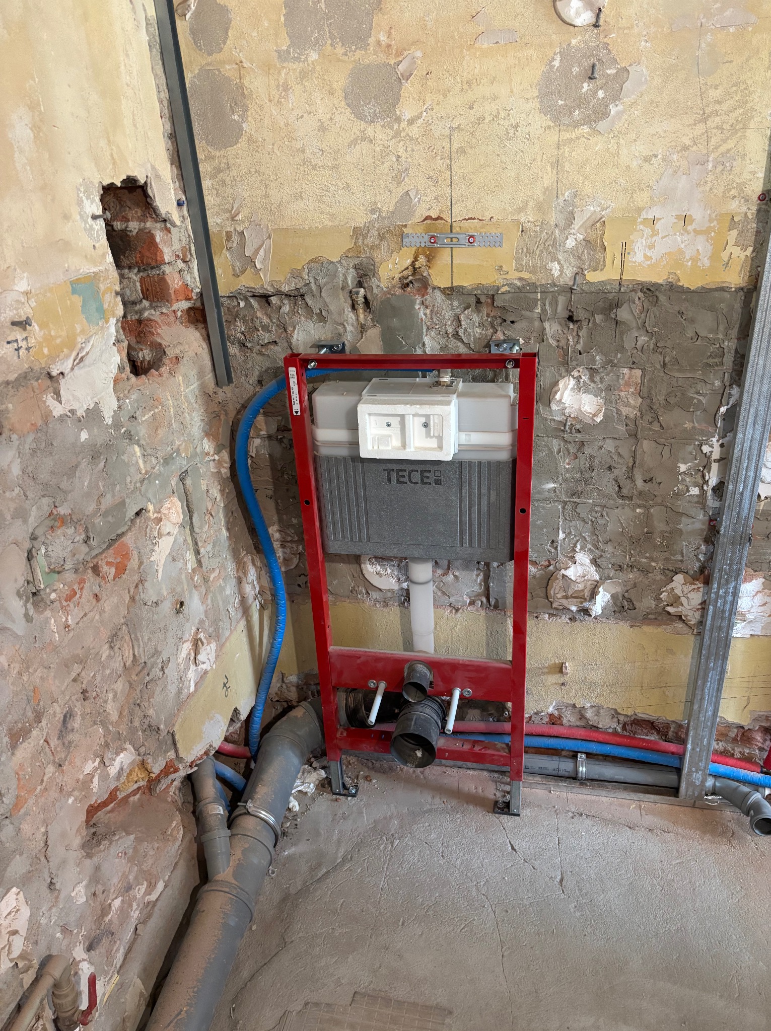 Stelaż podtynkowy TECE z instalacją wodno-kanalizacyjną na tle surowej ściany z cegłą i resztkami tynku, widoczne rury w kolorze niebieskim i czerwonym.