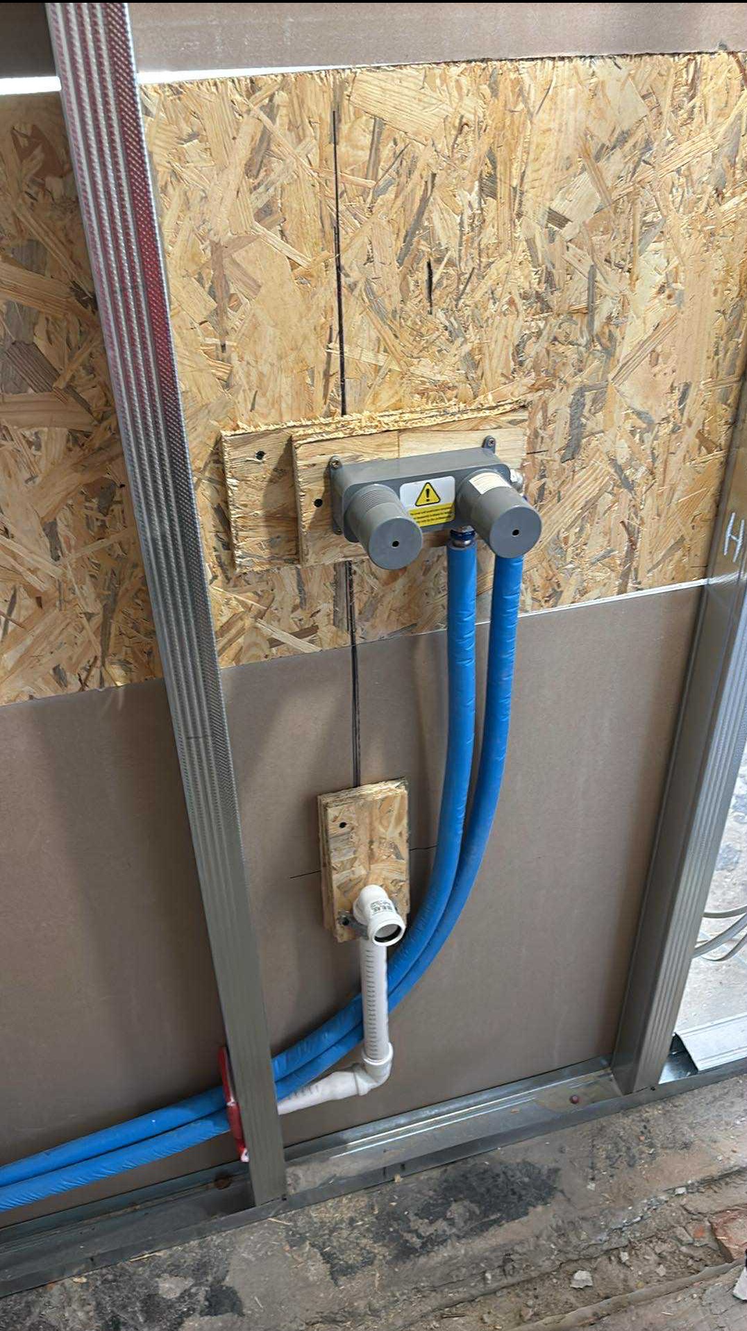 Fragment instalacji wodnej w ścianie z płyt OSB i metalowych profili, widoczne niebieskie rury PEX i szara skrzynka przyłączeniowa z symbolem ostrzegawczym.