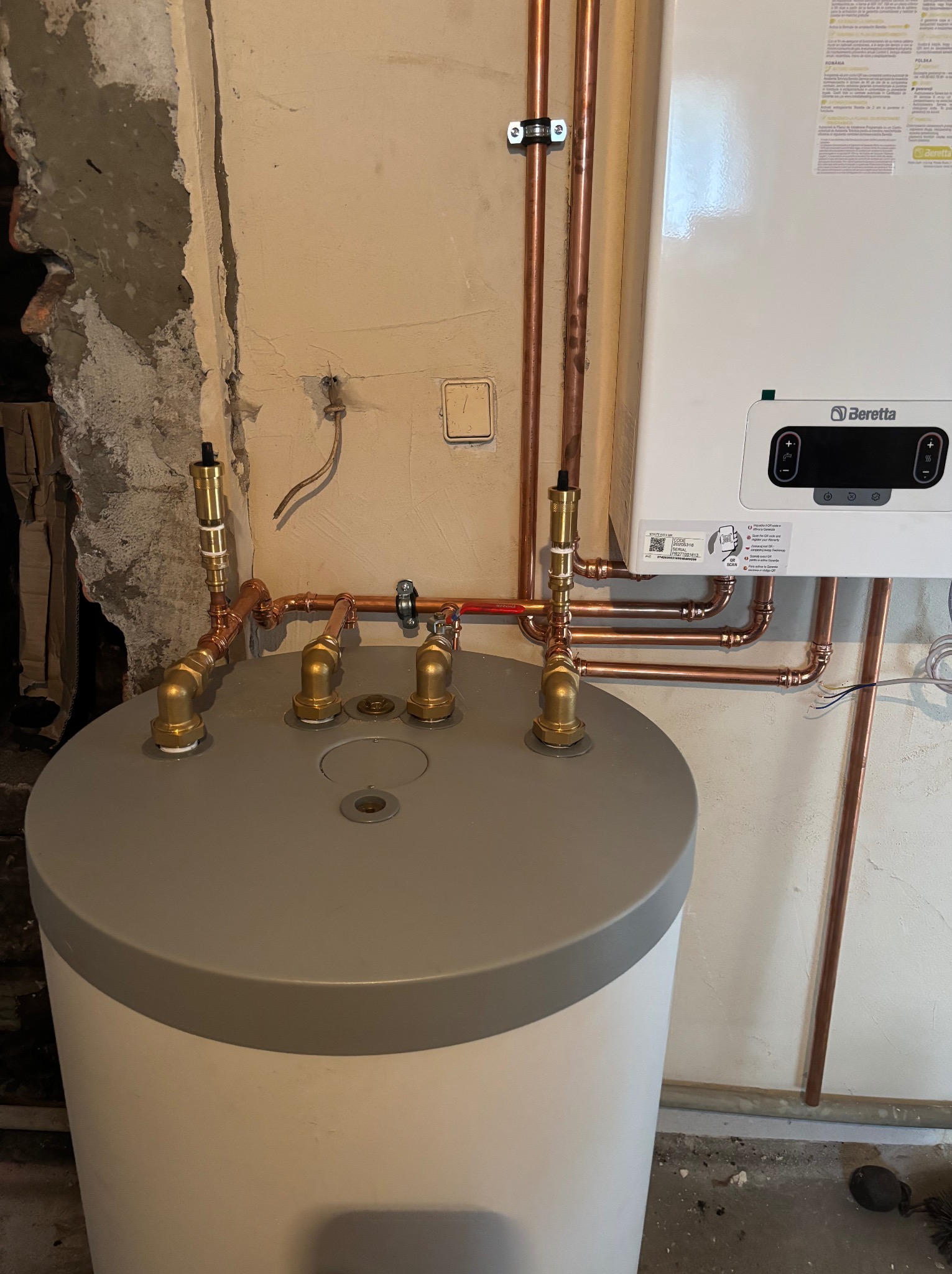 Nowy, biały zasobnik ciepłej wody użytkowej z miedzianą instalacją i piecem Beretta na ścianie. Widoczne łączenia i zawory. Surowa ściana w tle.