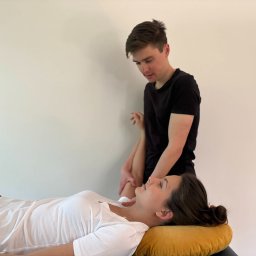 Fizjo Przestrzenie - Fizjoterapia Warszawa - Fizjoterapeuta masuje ramię pacjentki leżącej na żółtej poduszce na stole do masażu. Pacjentka w białej koszulce, fizjoterapeuta w czarnej.