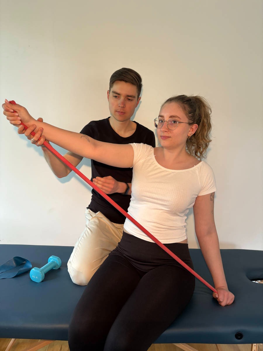 Kobieta w okularach ćwiczy z czerwoną gumą na kozetce, nadzorowana przez mężczyznę w czarnej koszulce. W tle ściana, obok leżą hantle.