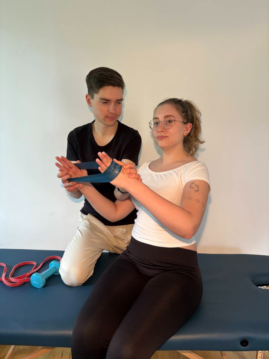 Sesja rehabilitacyjna: Mężczyzna pomaga kobiecie w ćwiczeniach z gumą na kozetce. W tle ściana, na kozetce leżą hantle i guma. Kobieta ma tatuaże na ramieniu.