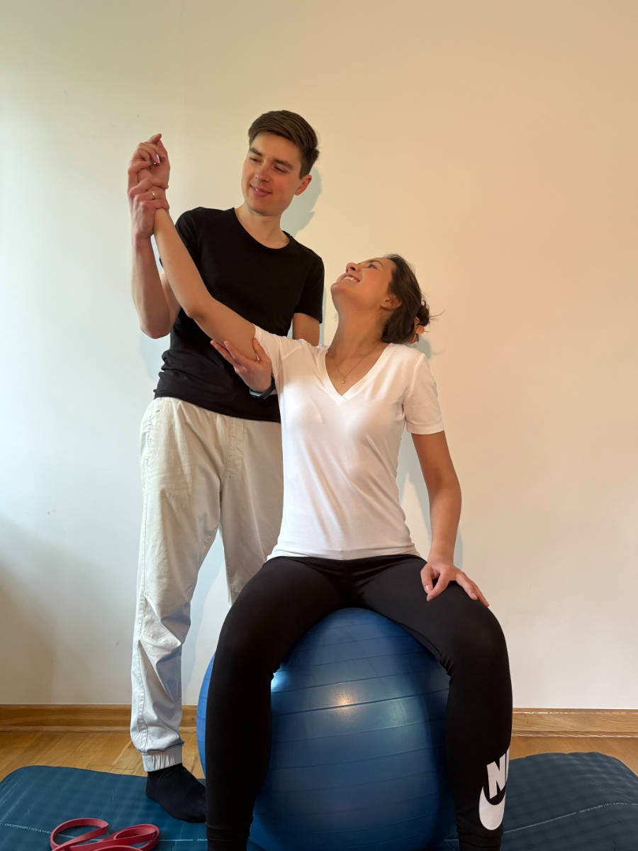 Mężczyzna wspiera kobietę siedzącą na piłce gimnastycznej podczas ćwiczeń rozciągających rękę, na macie do ćwiczeń leżą gumy oporowe.
