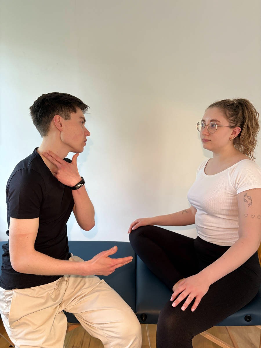 Fizjoterapeuta demonstruje pacjentce ćwiczenie na szyję. Pacjentka siedzi na krześle z nogami skrzyżowanymi. Białe tło, naturalne światło dzienne.