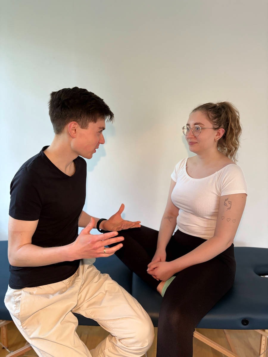 Konsultacja: mężczyzna w czarnym t-shircie wyjaśnia kobiecie w okularach siedzącej na leżance, gestykulując rękami. Jasne tło, naturalne światło.