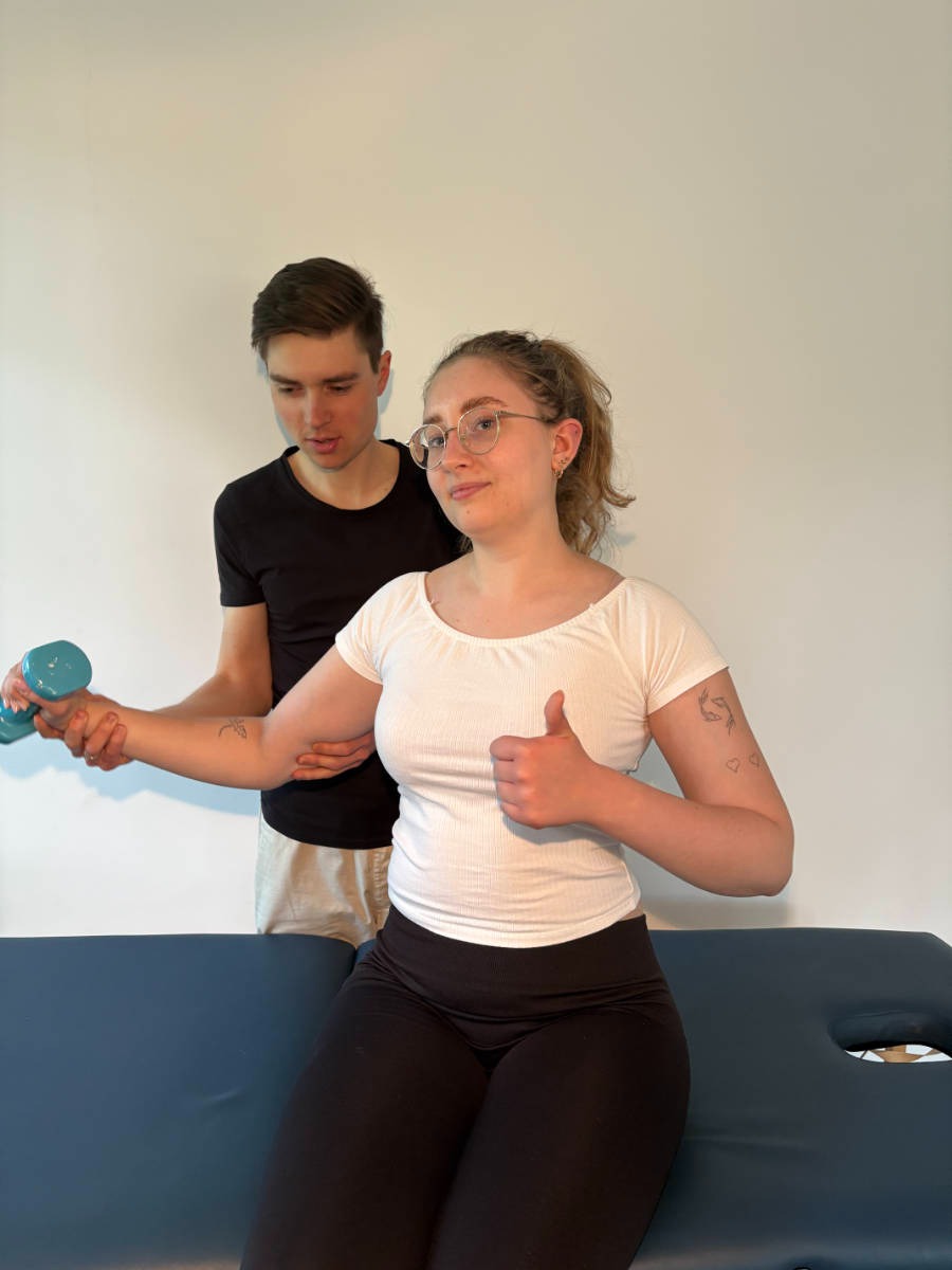 Pacjentka na kozetce ćwiczy z hantlą pod okiem mężczyzny; kciuk w górę, tatuaże na ramieniu, okulary w metalowej oprawce, czarne legginsy.