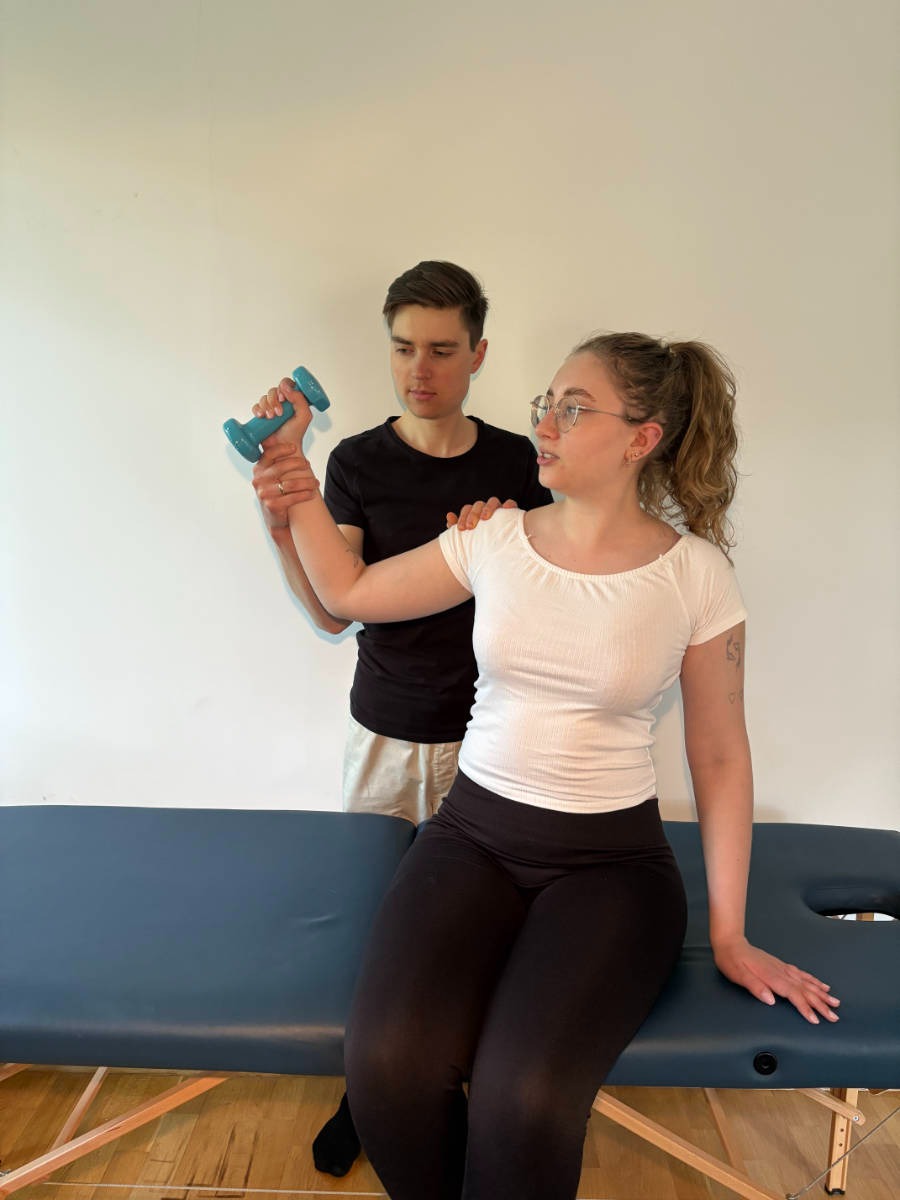 Kobieta ćwiczy z hantlą pod nadzorem mężczyzny na kozetce. Mężczyzna w czarnej koszulce, kobieta w białej bluzce i okularach. Jasne tło.