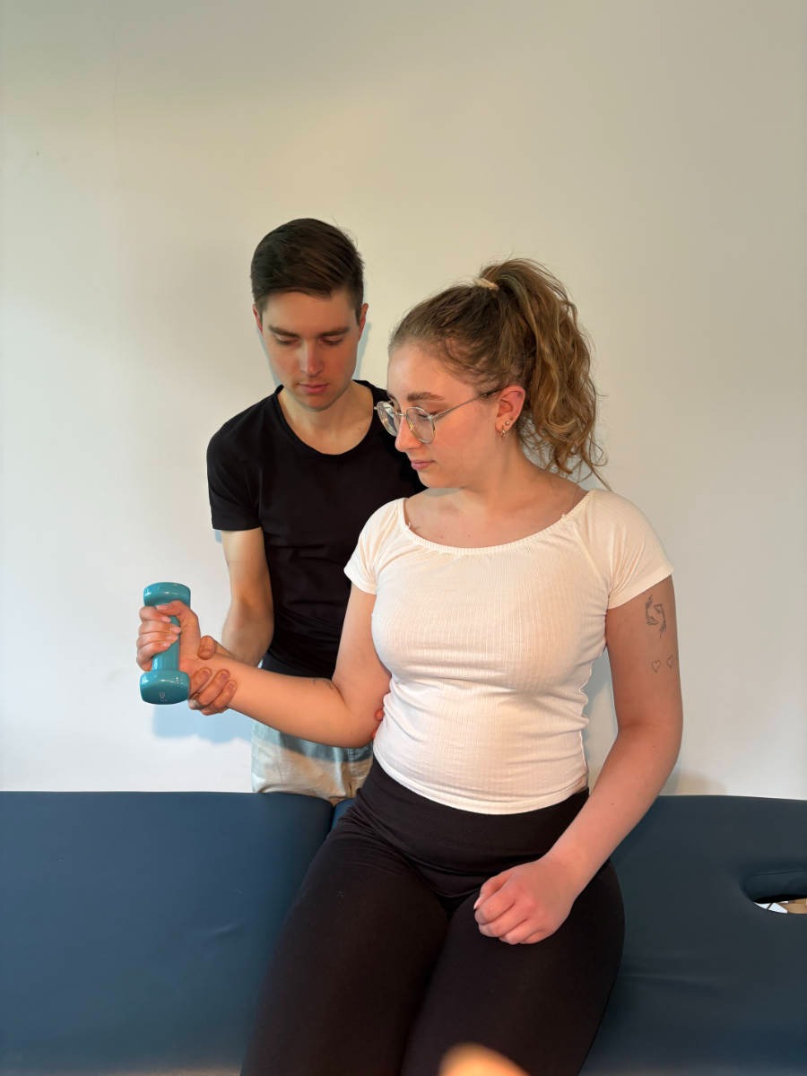 Kobieta w okularach ćwiczy z hantlą pod okiem mężczyzny na kozetce rehabilitacyjnej. Ćwiczenia wzmacniające pod nadzorem specjalisty.