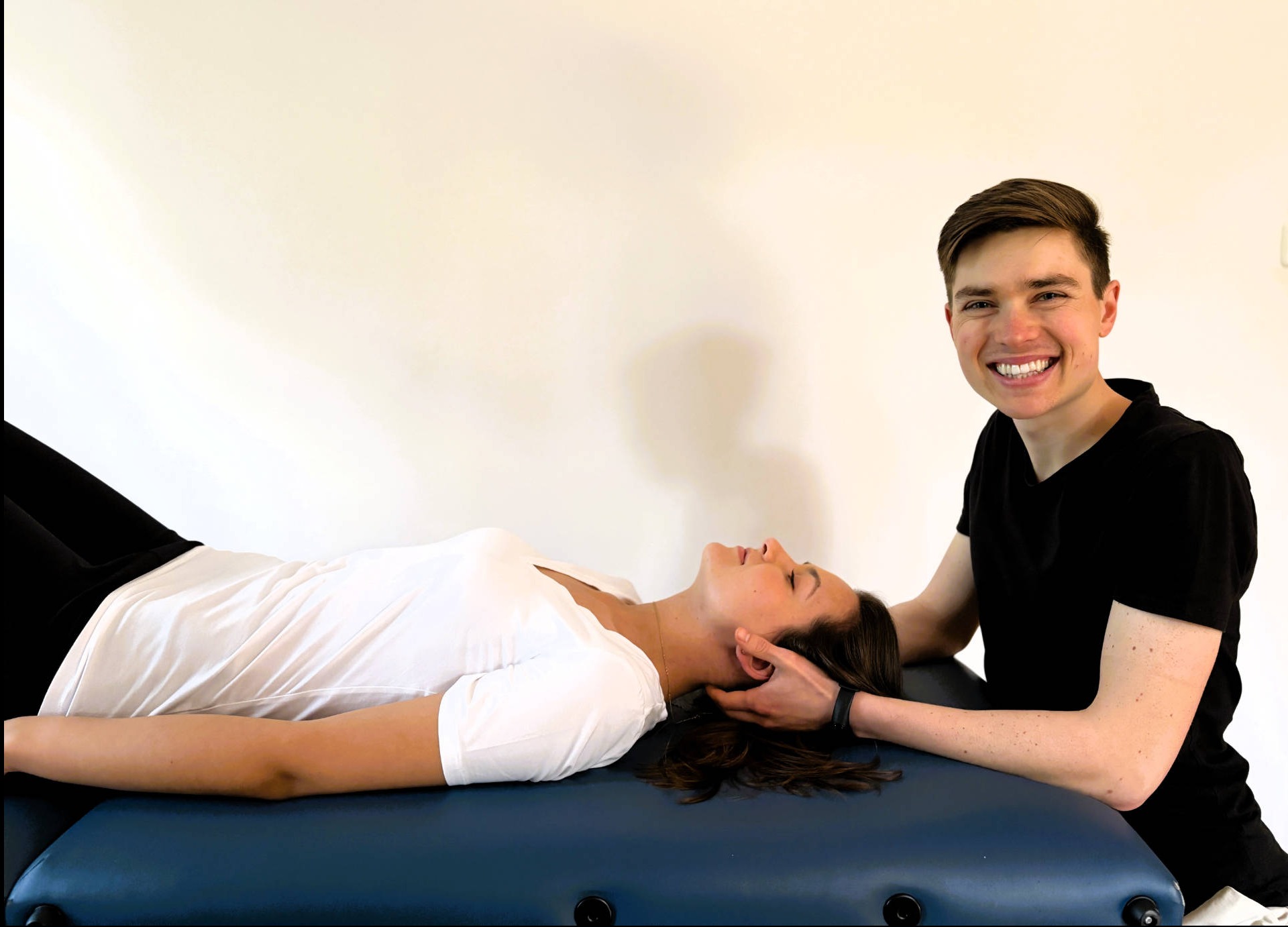 Kobieta leży na kozetce, a uśmiechnięty mężczyzna w czarnej koszulce wykonuje masaż szyi. Jasne tło, naturalne światło.