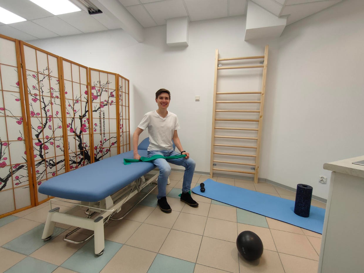 Uśmiechnięty mężczyzna siedzi na niebieskim stole rehabilitacyjnym, trzymając zieloną gumę do ćwiczeń. W tle drabinka gimnastyczna i mata do ćwiczeń.