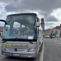 Paweł Barczykowski Usługi remontowo-budowlane. Transport osobowy - Srebrny autokar turystyczny Mercedes-Benz z żółtym napisem 'Barczykowski' na tle miejskiej architektury i pochmurnego nieba.