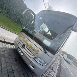 Paweł Barczykowski Usługi remontowo-budowlane. Transport osobowy - Szary autokar turystyczny z otwartymi drzwiami, zaparkowany na brukowanej ulicy, z zielonym żywopłotem w tle. Widoczny napis 'Barczykowski' na pojeździe.