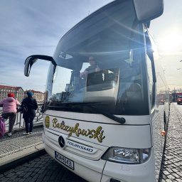 Paweł Barczykowski Usługi remontowo-budowlane. Transport osobowy - Biały autokar turystyczny z nazwą 'Barczykowski TOURISMO' stoi na brukowanej ulicy, a kierowca w okularach pokazuje kciuk w górę. W tle budynki i osoby.