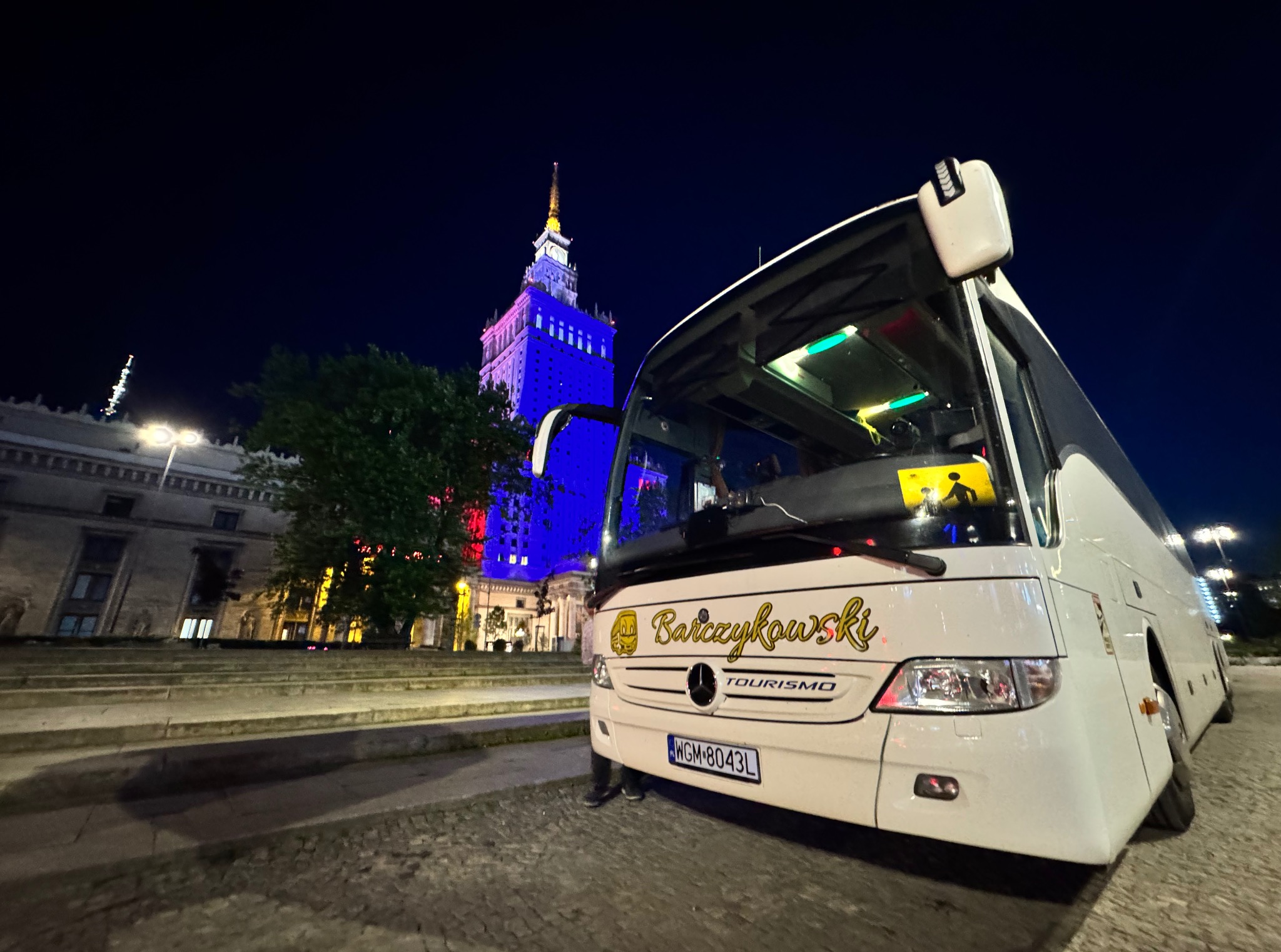 Biały autokar turystyczny z napisem 'Barczykowski' na tle nocnego krajobrazu Warszawy, w tle Pałac Kultury oświetlony na niebiesko i czerwono.
