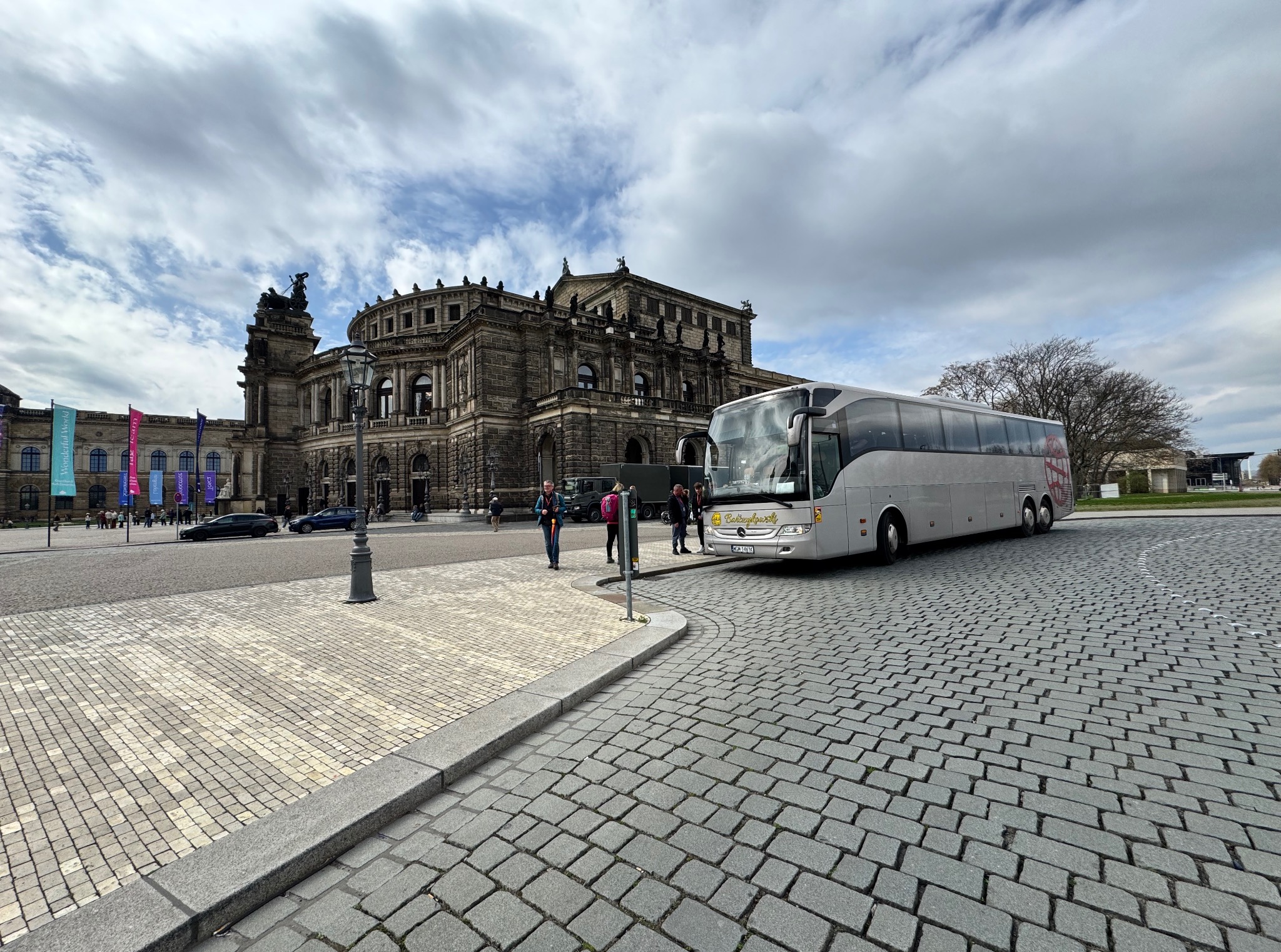 Jasny autokar turystyczny parkuje na brukowanym placu w Dreźnie, z Operą Semper w tle i przechodzącymi osobami. Pochmurne niebo.