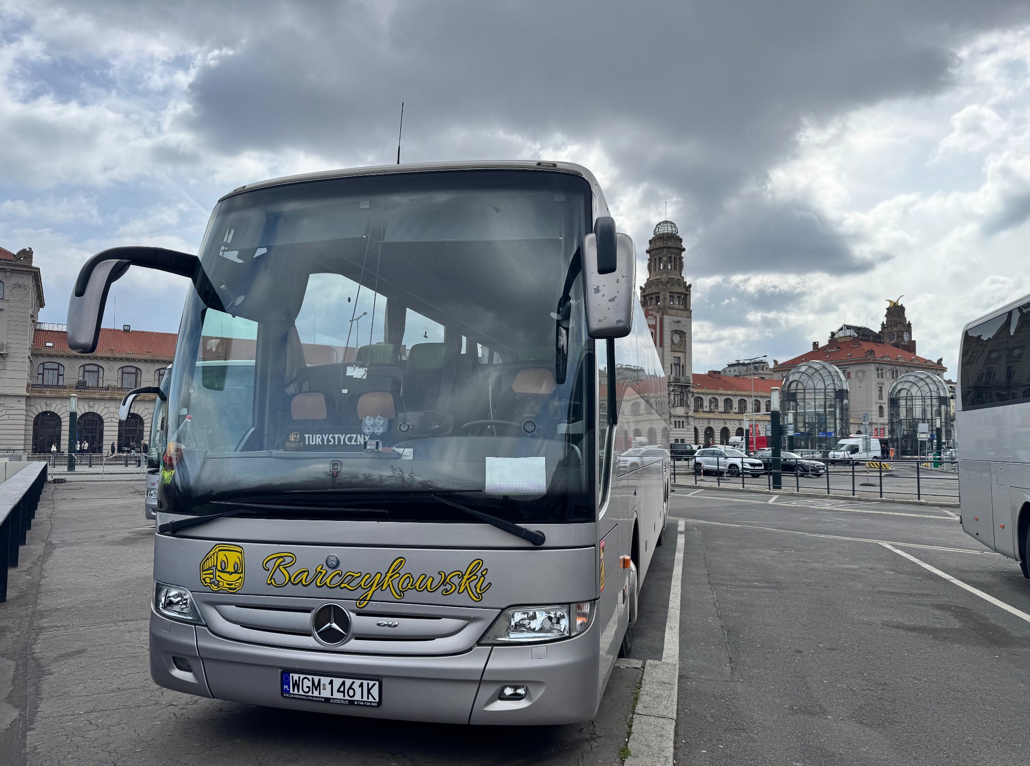 Srebrny autokar turystyczny Mercedes-Benz z żółtym napisem 'Barczykowski' na tle miejskiej architektury i pochmurnego nieba.