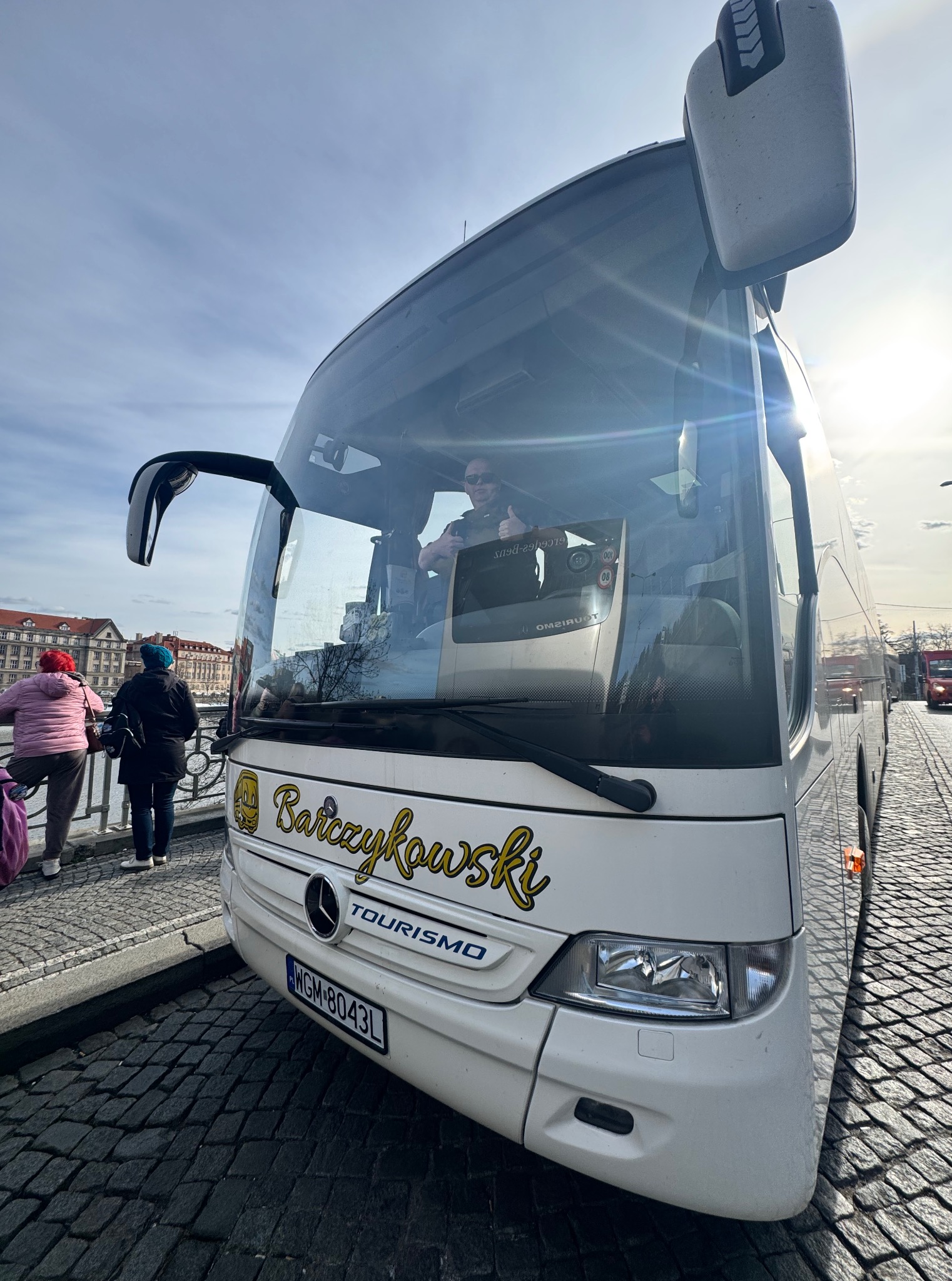 Biały autokar turystyczny z nazwą 'Barczykowski TOURISMO' stoi na brukowanej ulicy, a kierowca w okularach pokazuje kciuk w górę. W tle budynki i osoby.