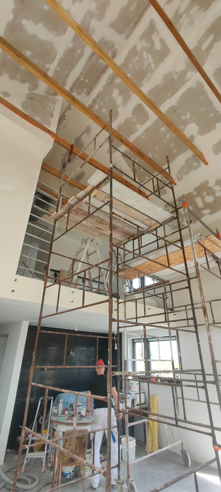 Wnętrze w trakcie prac wykończeniowych: sufit z płyt karton-gips, drewniane belki i rusztowanie. Widoczne narzędzia i materiały budowlane.