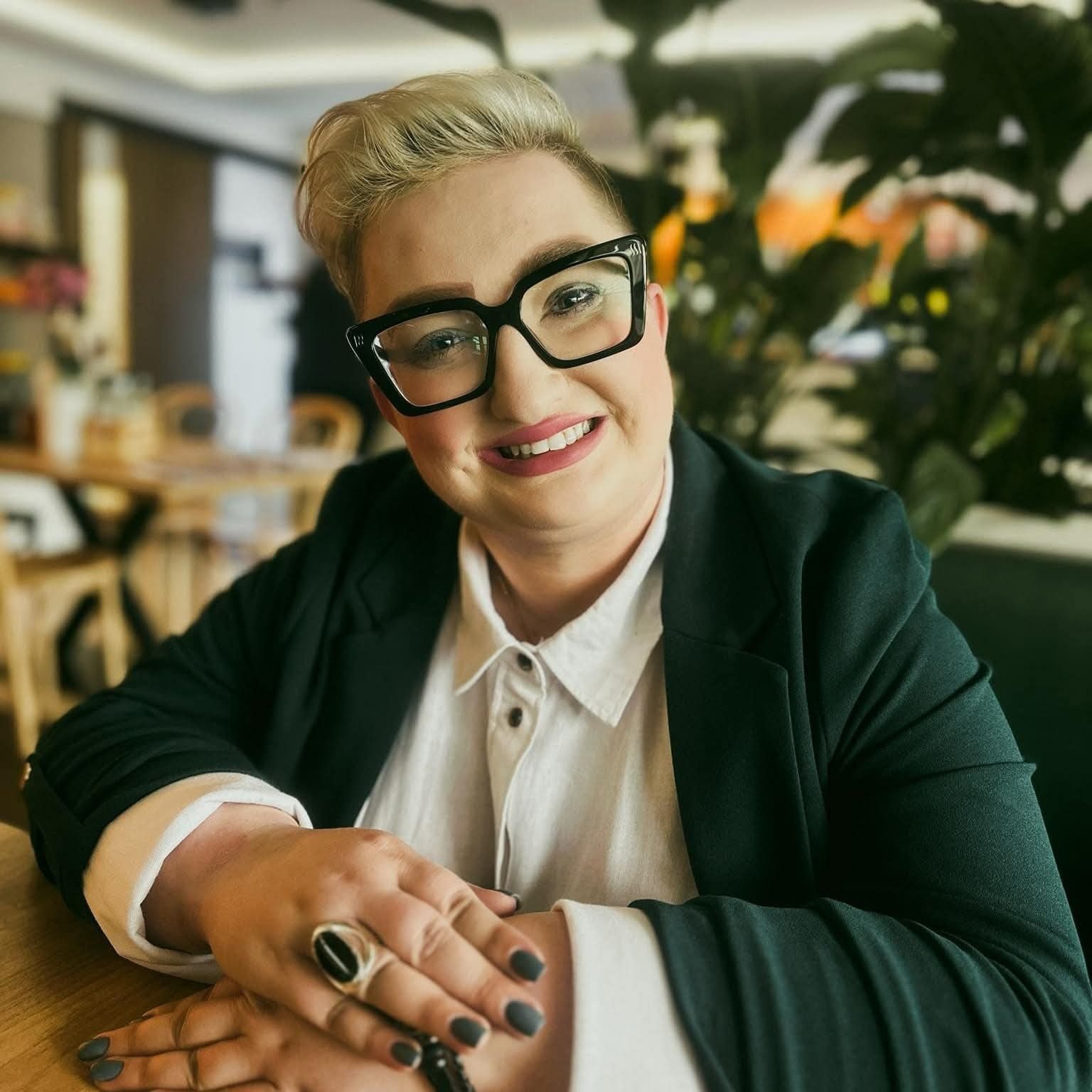 Uśmiechnięta kobieta w okularach i ciemnozielonej marynarce, siedząca przy drewnianym stole z dłońmi złożonymi przed sobą, w tle widoczna roślina doniczkowa.