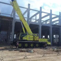 Żółty dźwig KAMATRANS na budowie hali z betonu, widoczne rusztowania i barierki ochronne. Błękitne niebo z chmurami w tle.