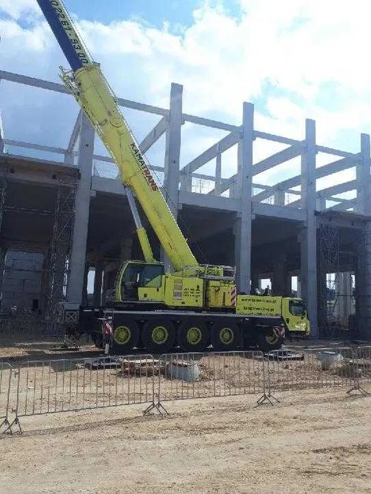 Żółty dźwig KAMATRANS na budowie hali z betonu, widoczne rusztowania i barierki ochronne. Błękitne niebo z chmurami w tle.