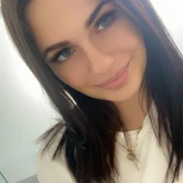 Portret uśmiechniętej brunetki z delikatnym makijażem i złotym naszyjnikiem na białym tle. Widoczne długie rzęsy i subtelny uśmiech.