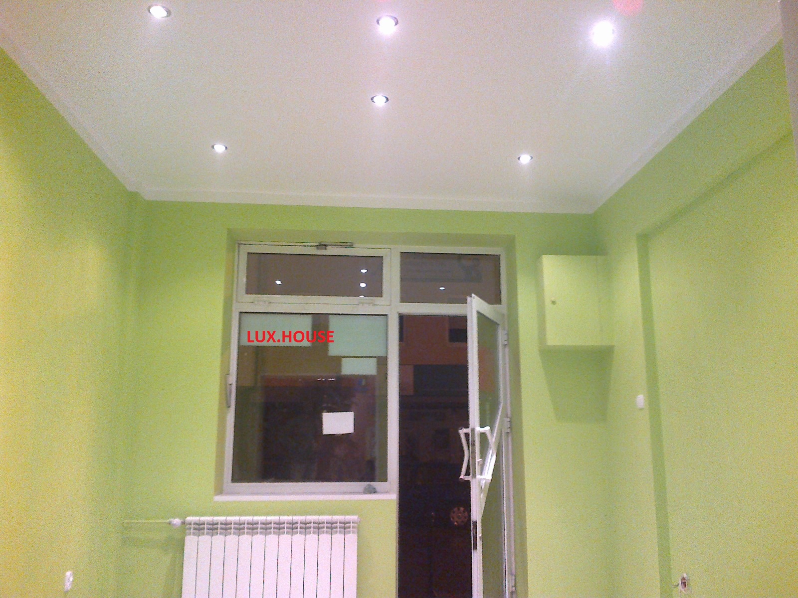 Pomalowane na jasnozielono wnętrze pokoju z białym sufitem i listwami, widoczne okno z napisem LUX.HOUSE, biały grzejnik i otwarte drzwi.