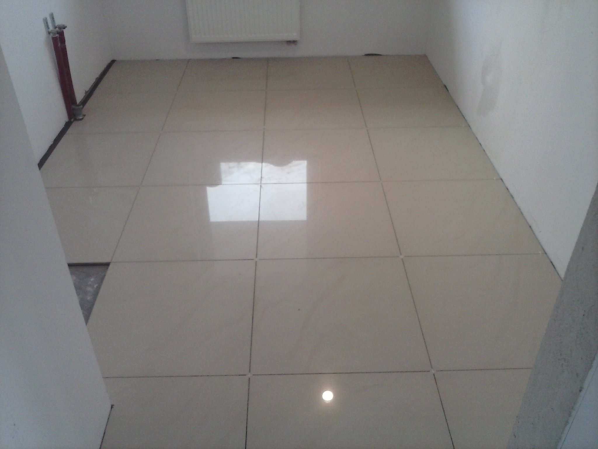 Jasne, kwadratowe płytki podłogowe z fugami, częściowo ułożone w pomieszczeniu z grzejnikiem i instalacją hydrauliczną przy ścianie, widoczny fragment nieukończonej podłogi.