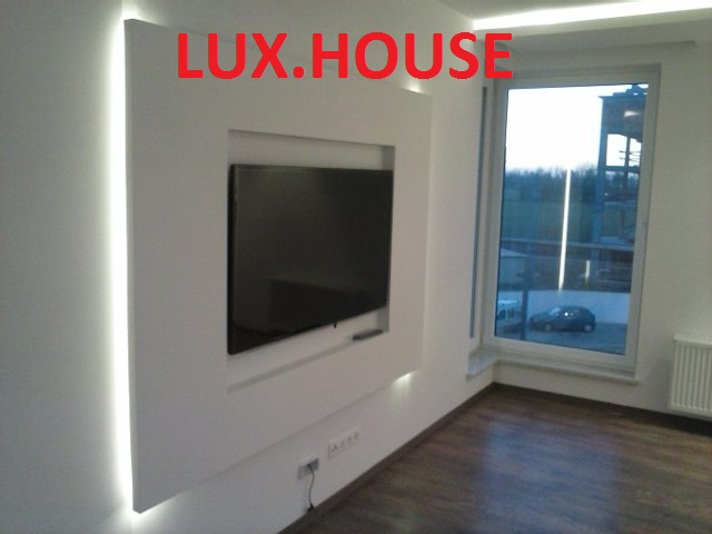 Nowoczesna zabudowa ściany z podświetleniem LED wokół wnęki na telewizor, widoczne gniazdka elektryczne i drewnopodobna podłoga.