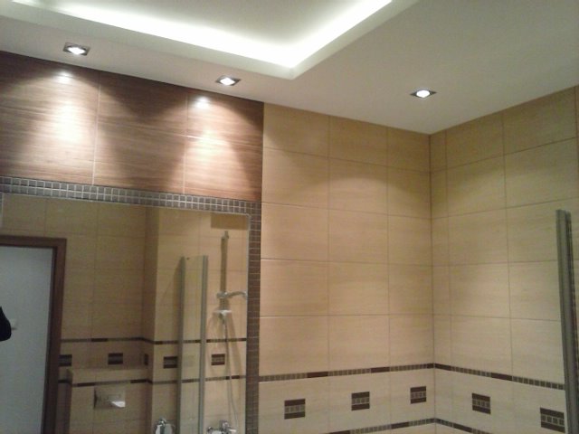 Wykończona łazienka z beżowymi i brązowymi płytkami, oświetleniem punktowym w suficie i lustrem z widoczną armaturą.