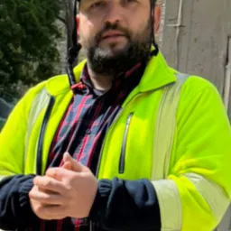 Mężczyzna w kasku BHP z lampką czołową, w neonowej kurtce i kraciastej koszuli, z zaciśniętymi dłońmi na tle drzew i betonowej konstrukcji.