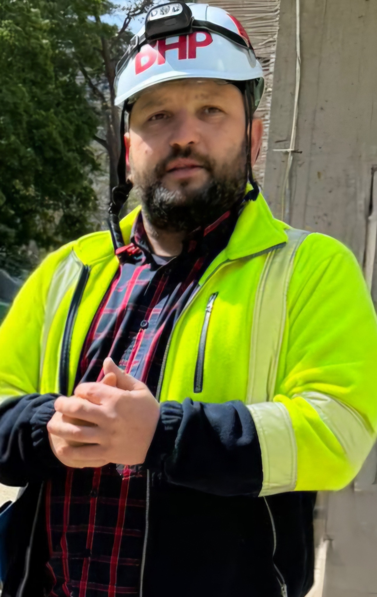 Mężczyzna w kasku BHP z lampką czołową, w neonowej kurtce i kraciastej koszuli, z zaciśniętymi dłońmi na tle drzew i betonowej konstrukcji.