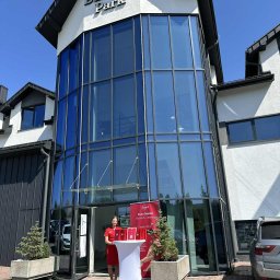 DeepilStudio Depilacja Rabka-Zdrój - Budynek Bulwary Park. Przed wejściem kobieta w czerwonej sukience i stoisko z materiałami reklamowymi. Promocja studia depilacji w Rabce-Zdroju.