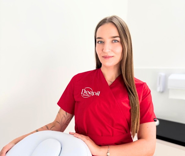Kobieta w czerwonym uniformie Deepil Studio, z delikatnym tatuażem na przedramieniu, pozuje w gabinecie kosmetycznym. Widoczny fotel zabiegowy.