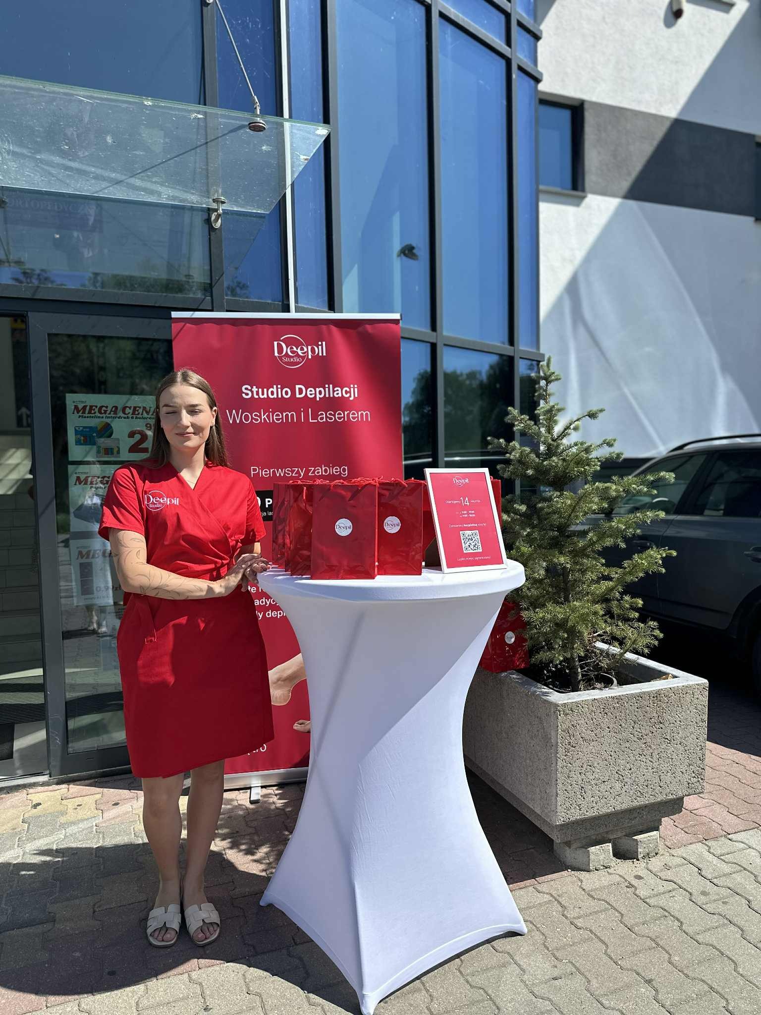 Promocja studia depilacji: kobieta w czerwonym uniformie, stół z upominkami, baner reklamowy i choinka przed wejściem do salonu. Widoczny kod QR.