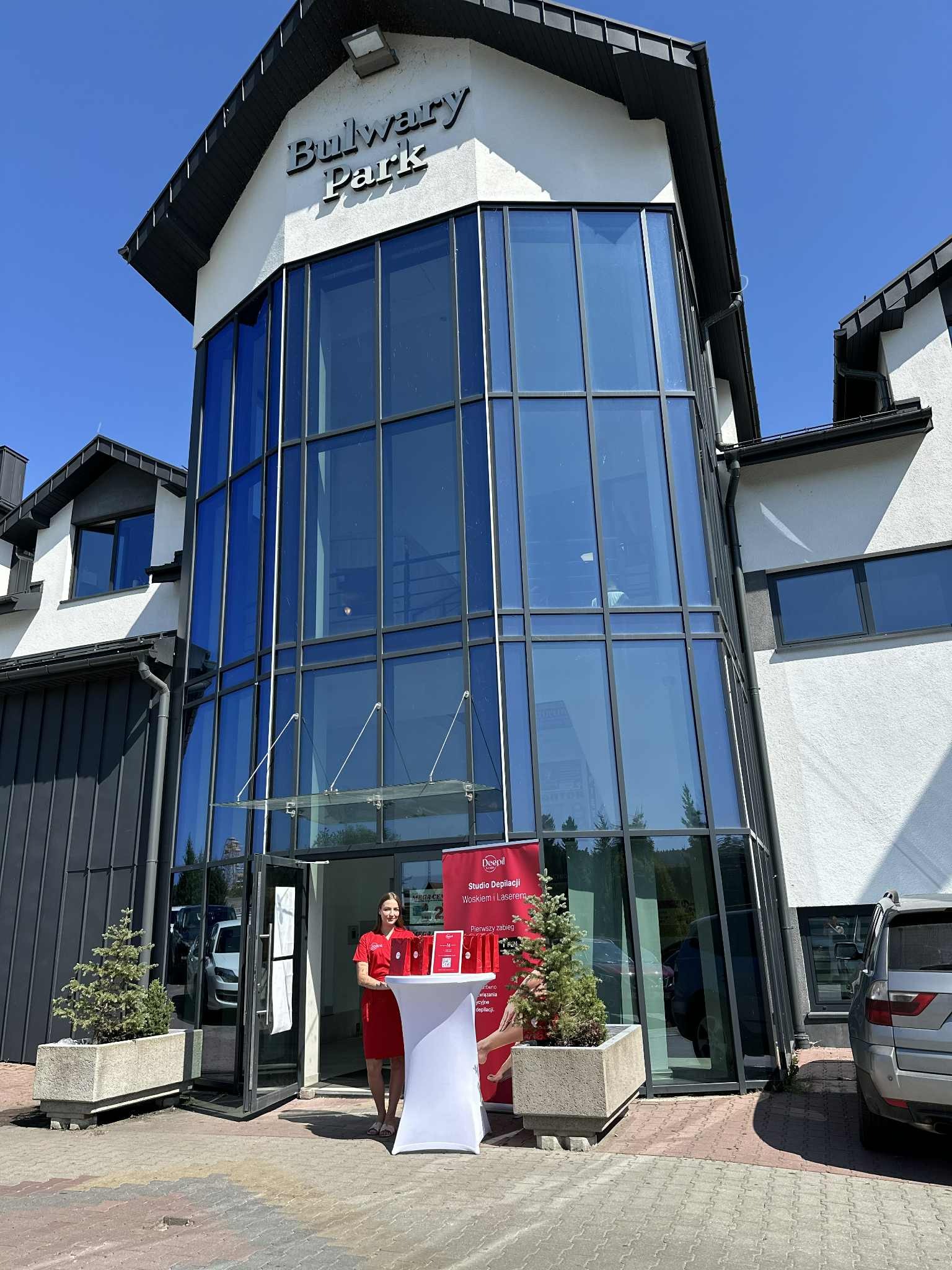 Budynek Bulwary Park. Przed wejściem kobieta w czerwonej sukience i stoisko z materiałami reklamowymi. Promocja studia depilacji w Rabce-Zdroju.