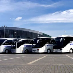 Flota autokarów Orland na tle nowoczesnej hali widowiskowej pod błękitnym niebem. Autokary w kolorach bieli i czerni, z logo firmy.