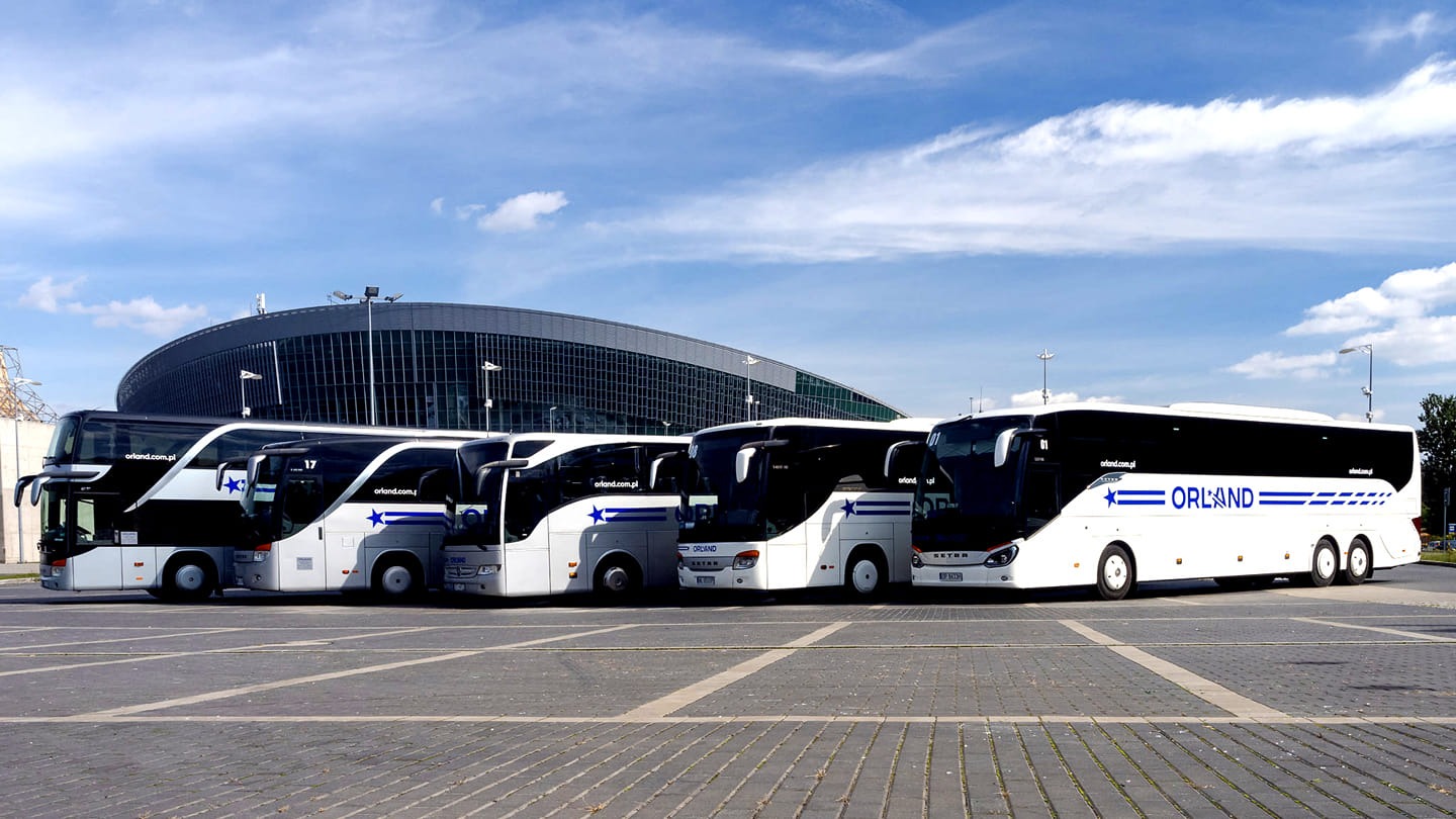 Flota autokarów Orland na tle nowoczesnej hali widowiskowej pod błękitnym niebem. Autokary w kolorach bieli i czerni, z logo firmy.