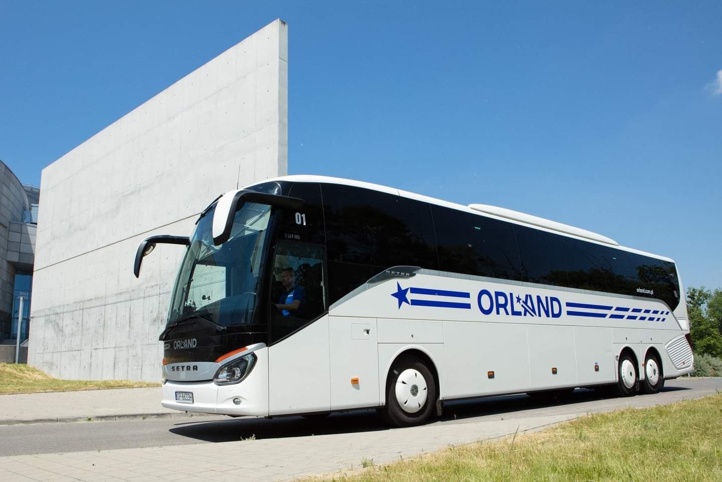 Biały autokar marki Setra z niebieskim logo Orland, zaparkowany na tle nowoczesnego budynku i błękitnego nieba. Widoczny kierowca w kabinie.