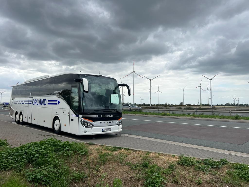 Biały autokar marki Setra z logo Orland na tle pochmurnego nieba i turbin wiatrowych, zaparkowany przy drodze. Widoczny fragment zieleni i chodnika.