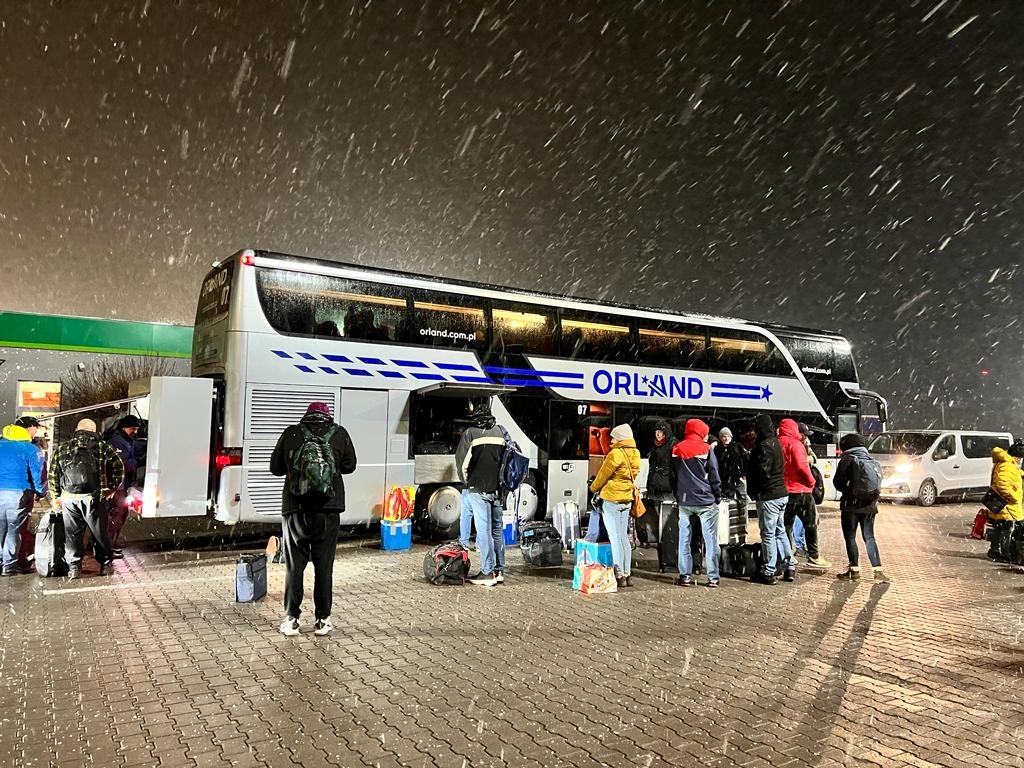 Autokar z pasażerami w zimowej scenerii. Osoby z bagażami wsiadają do pojazdu marki Orland podczas śnieżycy. Widoczny biały bus w tle.