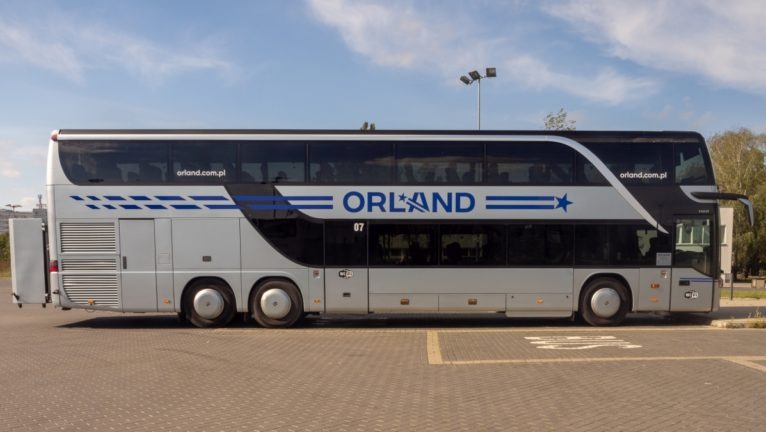Dwupiętrowy autobus firmy Orland z Żędowic, widok z boku na tle błękitnego nieba. Autobus w kolorze srebrnym z niebieskimi akcentami i logo firmy.